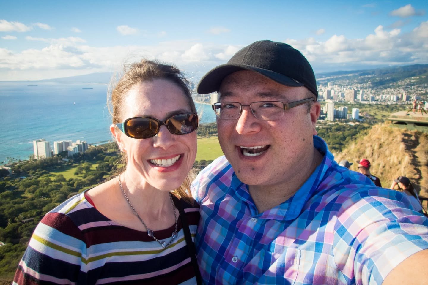 Oahu Two travelers taking a selfie at a scenic overlook, with the coastline and an urban cityscape visible in the background.