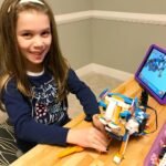 Oahu A child smiles while working with a colorful buildable toy and a tablet on a wooden table.