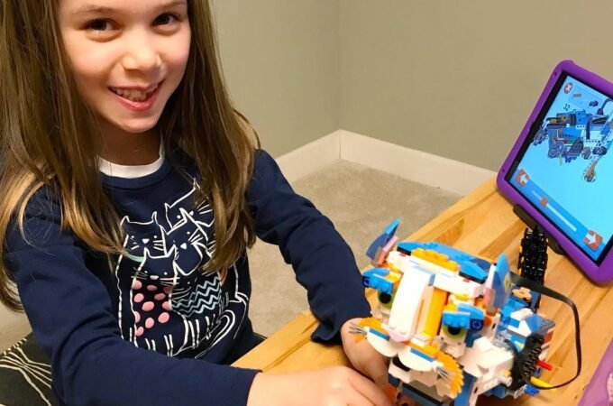 A child smiles while working with a colorful buildable toy and a tablet on a wooden table.