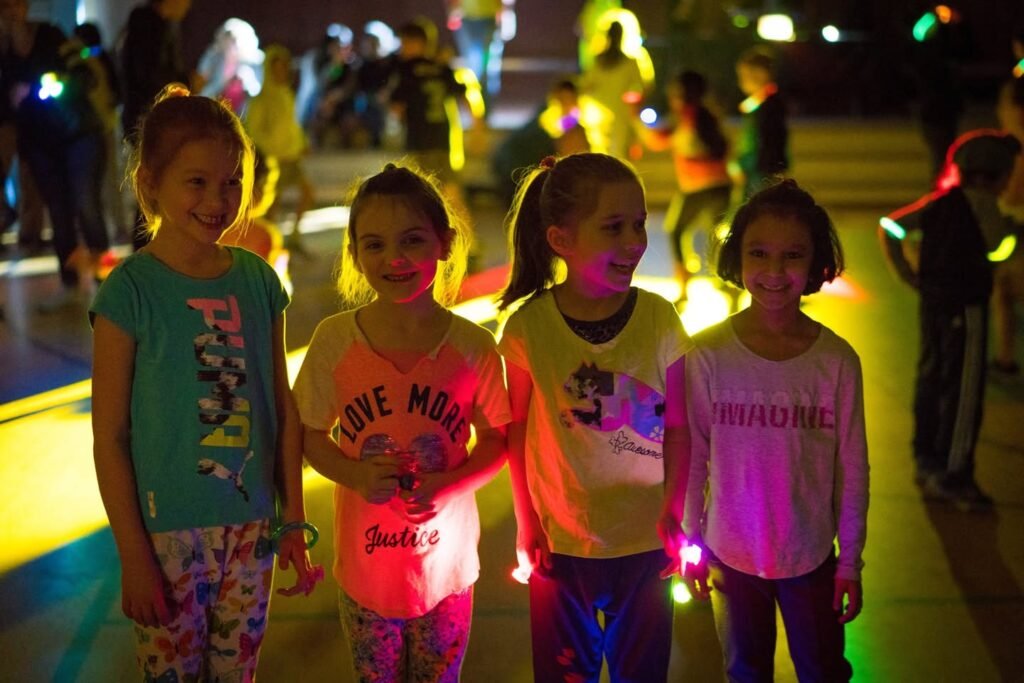 Young girls wearing glow bands and shirts at a colored light event, smiling for a photo at night.