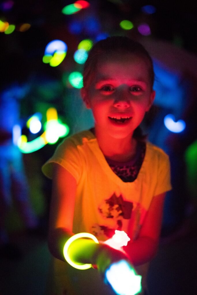 A smiling child wearing a yellow shirt holds glowing toys amidst vibrant, bokeh lights in a dark setting.