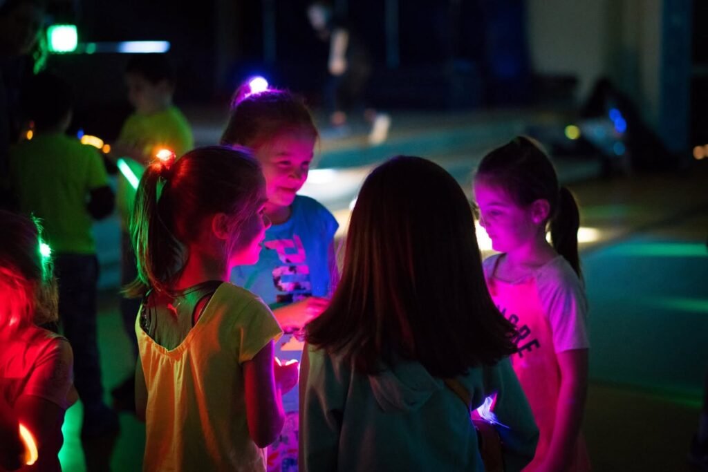 Children wearing glow sticks gather in the dark at a nighttime neighborhood event.