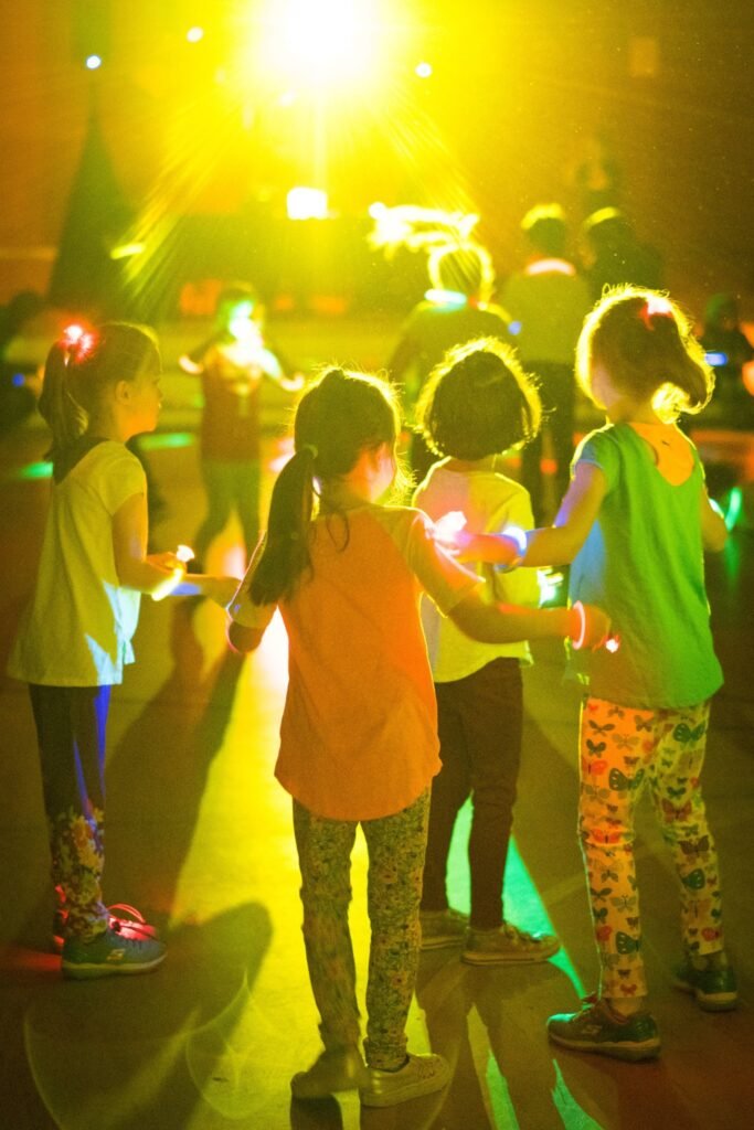 A group of children hold hands and dance under bright, colorful stage lights at an event.