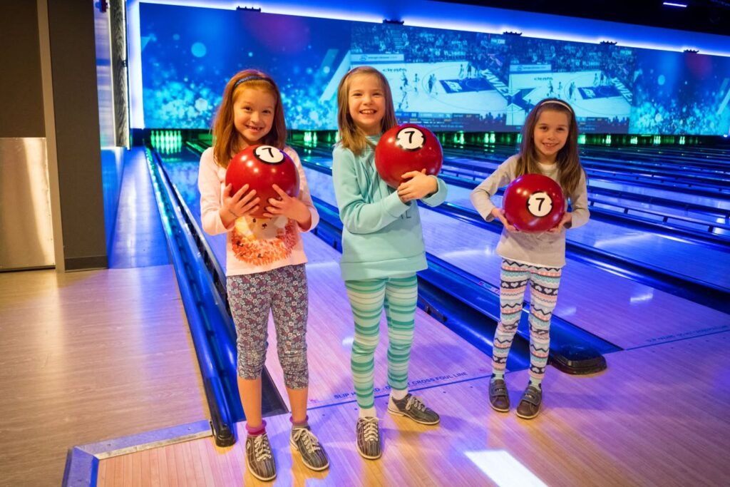 Three girls standing at the end of a bowling lane holding red balls, with a vibrant blue digital display in the background.
