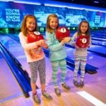 Kids holding red bowling balls in a modern bowling alley with a large screen displaying a motivational quote.