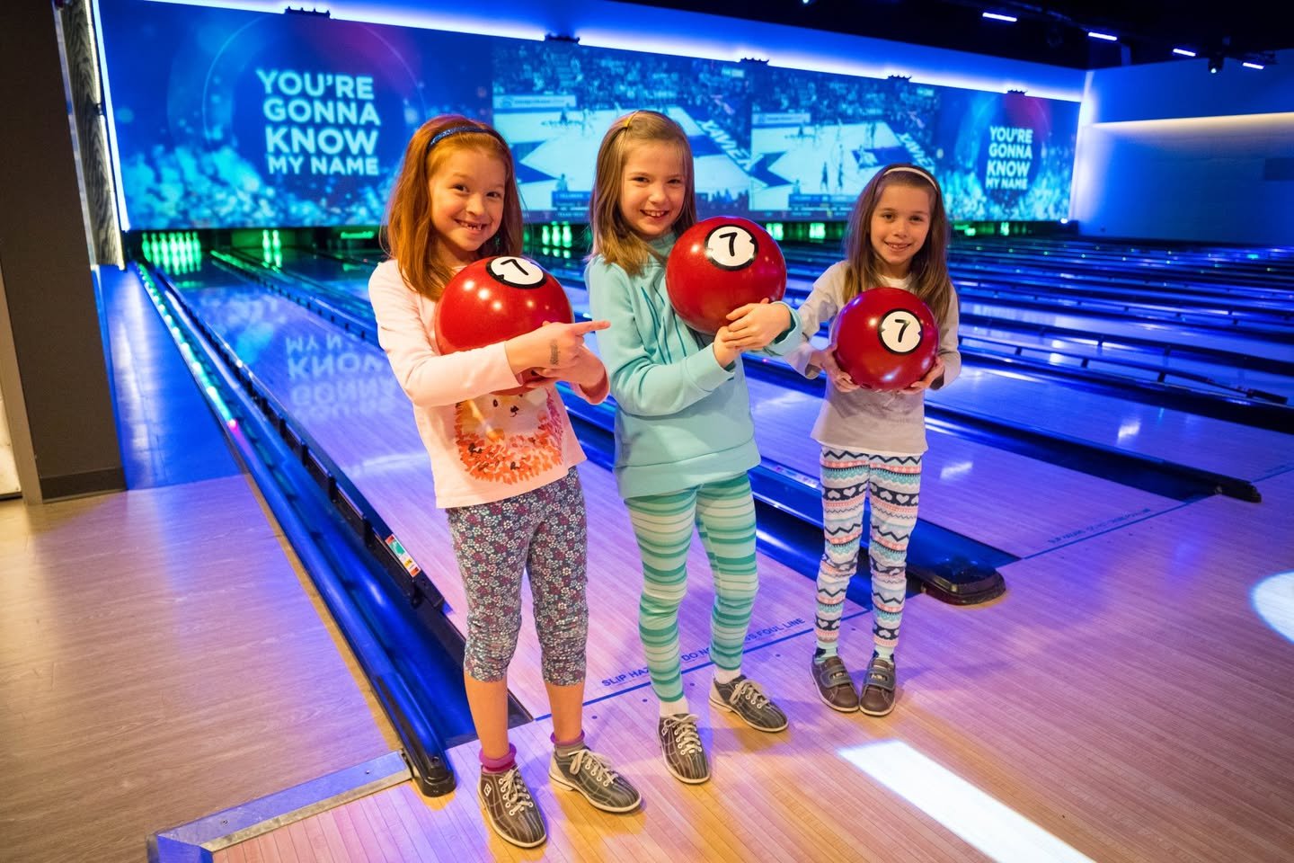 Kids holding red bowling balls in a modern bowling alley with a large screen displaying a motivational quote.
