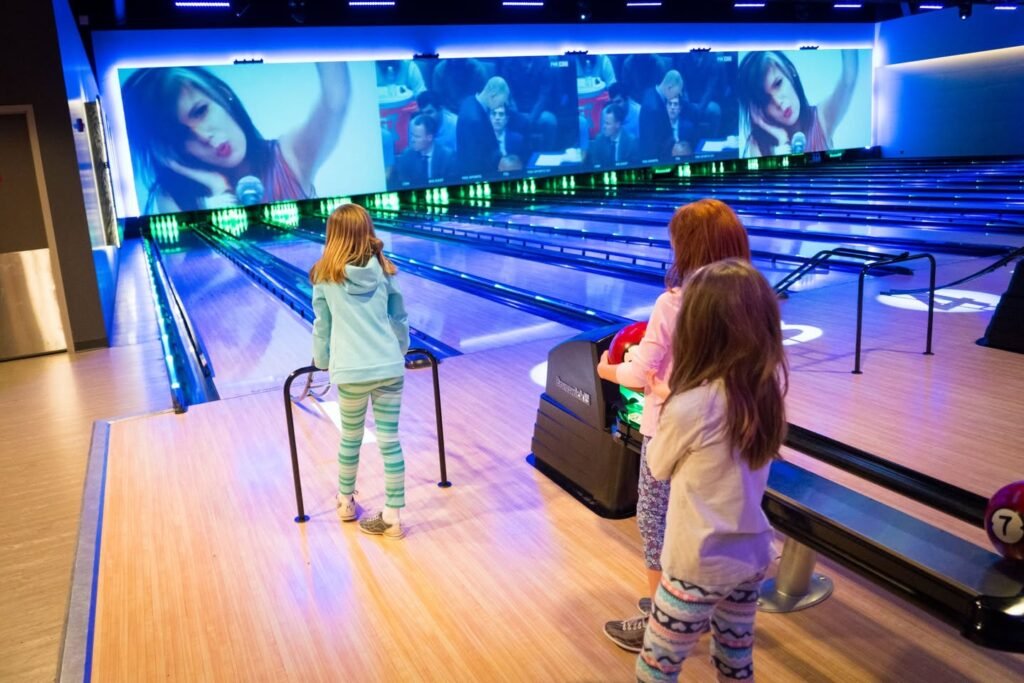 Children enjoying a game at a modern bowling alley with immersive digital displays. The lanes are lit with dynamic blue and green lights.