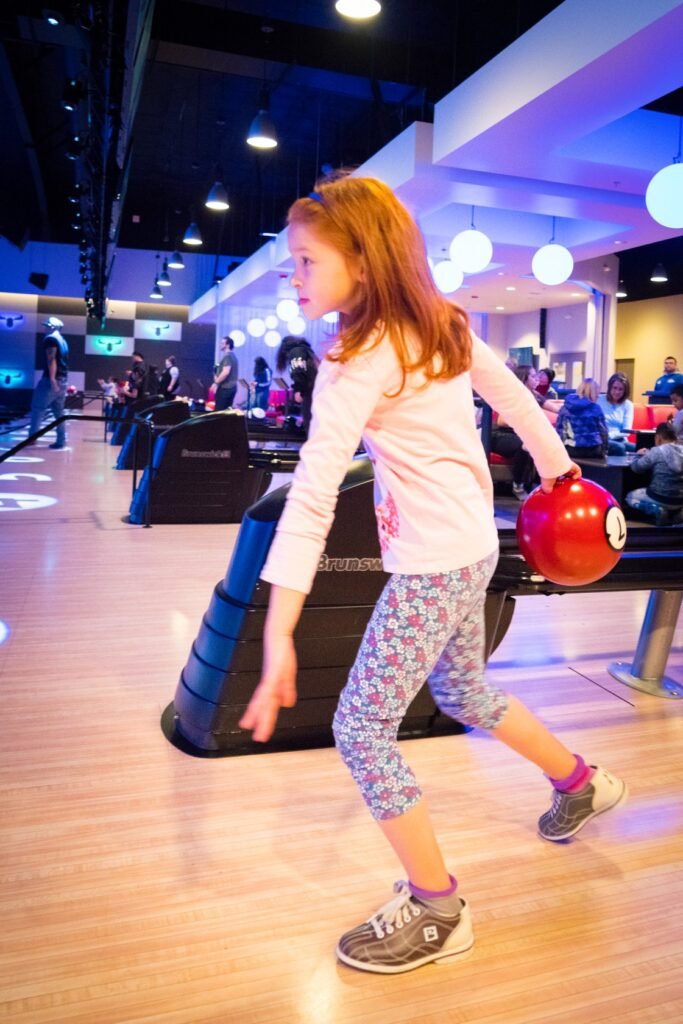 A young girl with reddish hair is holding red bowling balls in a bowling alley.