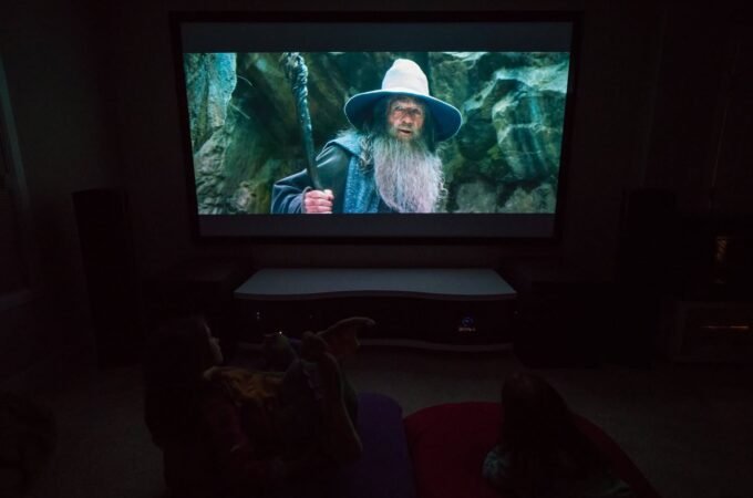 A dark home theater with a projector displaying a scene from a fantasy film featuring a wizard in a wide-brimmed hat.