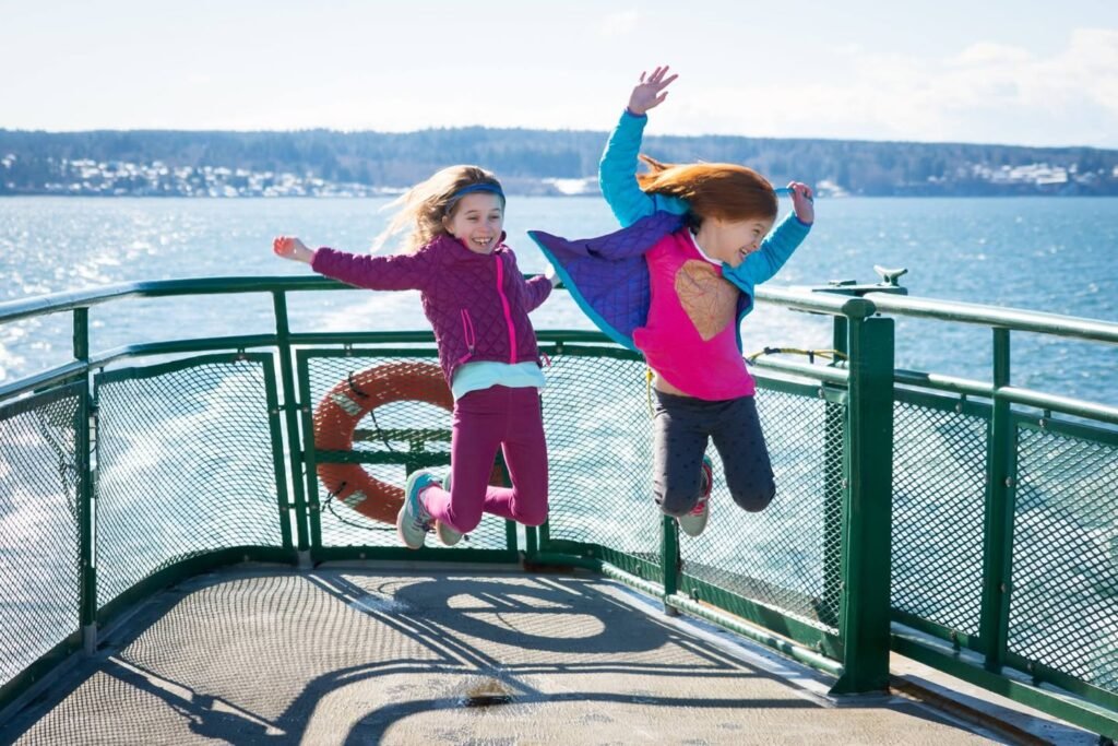 Two children jumping and running along the outdoor promenade of a ferry boat.
