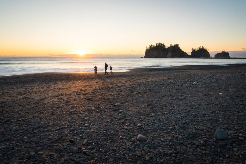 A sun setting over the ocean with rocky outcroppings in the distance. Several people are walking along the shoreline on a coastal beach.