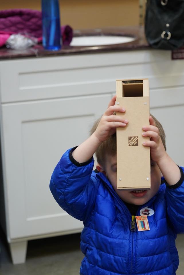 Child in a blue jacket holding a tall wooden block over their face. A smaller close-up of a tote bag and countertop.