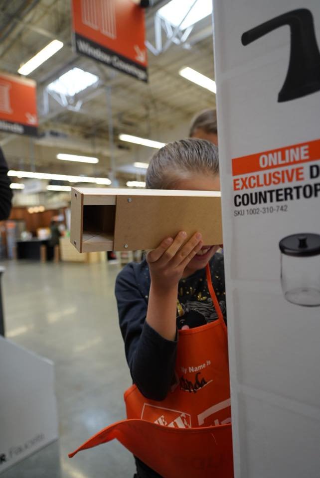 A woman wearing an orange apron hides behind a display with a cardboard box while a child peeks from behind it in a home improvement store.
