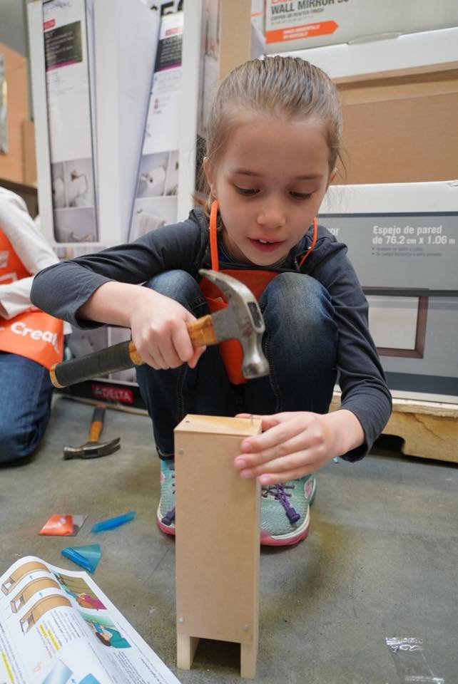 A person wearing a safety vest is squatting on the floor and using a hammer to nail a small wooden structure.