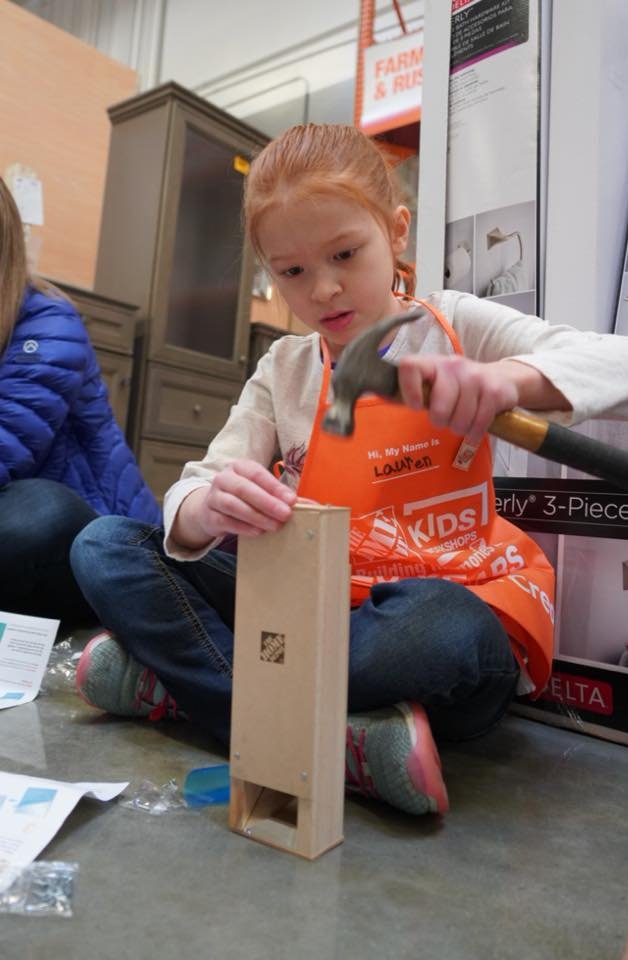 A child sits on the floor assembling a small piece of furniture while wearing an orange apron. Another person in a blue jacket is nearby.