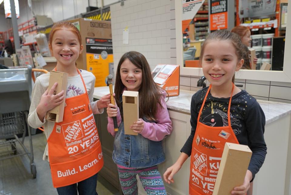 Three children wearing orange aprons holding wooden boards at Home Depot. Another child wearing an apron is shown smiling with a close-up of the apron's 20th anniversary design.