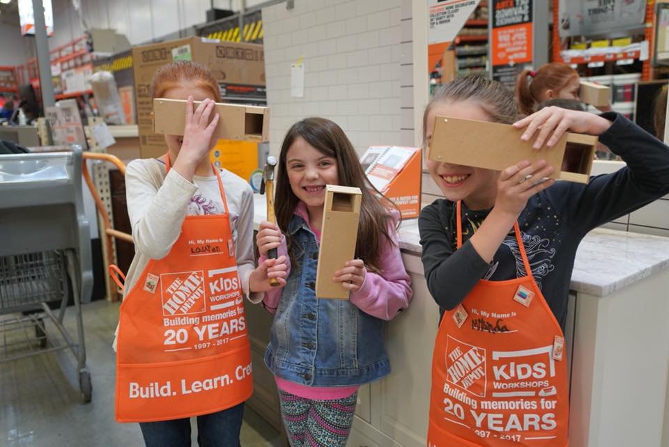 Children wearing orange Home Depot aprons hold wooden objects inside a retail store.