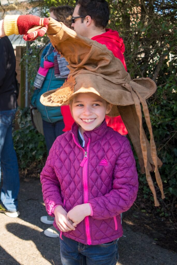 A smiling child in a purple quilted jacket and hat posing outdoors with others in a garden setting. Another person is seen holding a brown fabric element.