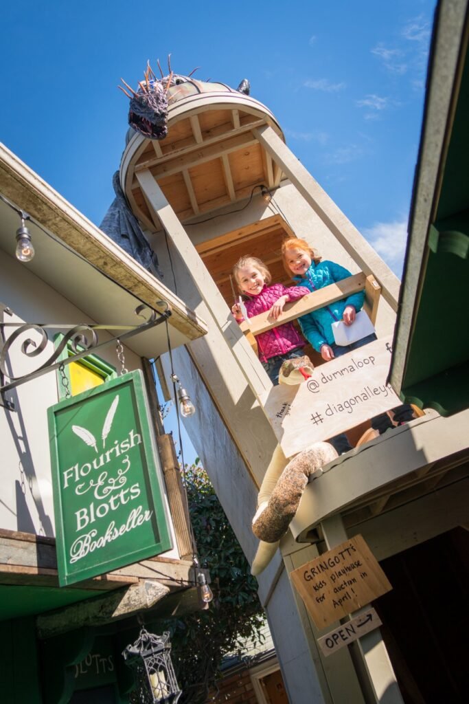 A flock of pigeons nests on a ledge above a storefront window where people stand, with a green and white sign attached to the building facade.