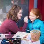 A scene of face painting at an indoor craft activity. An individual is applying paint to another's face in a colorful setting.