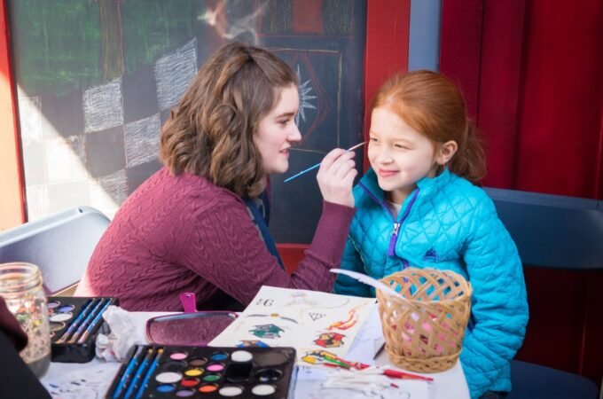 A scene of face painting at an indoor craft activity. An individual is applying paint to another's face in a colorful setting.
