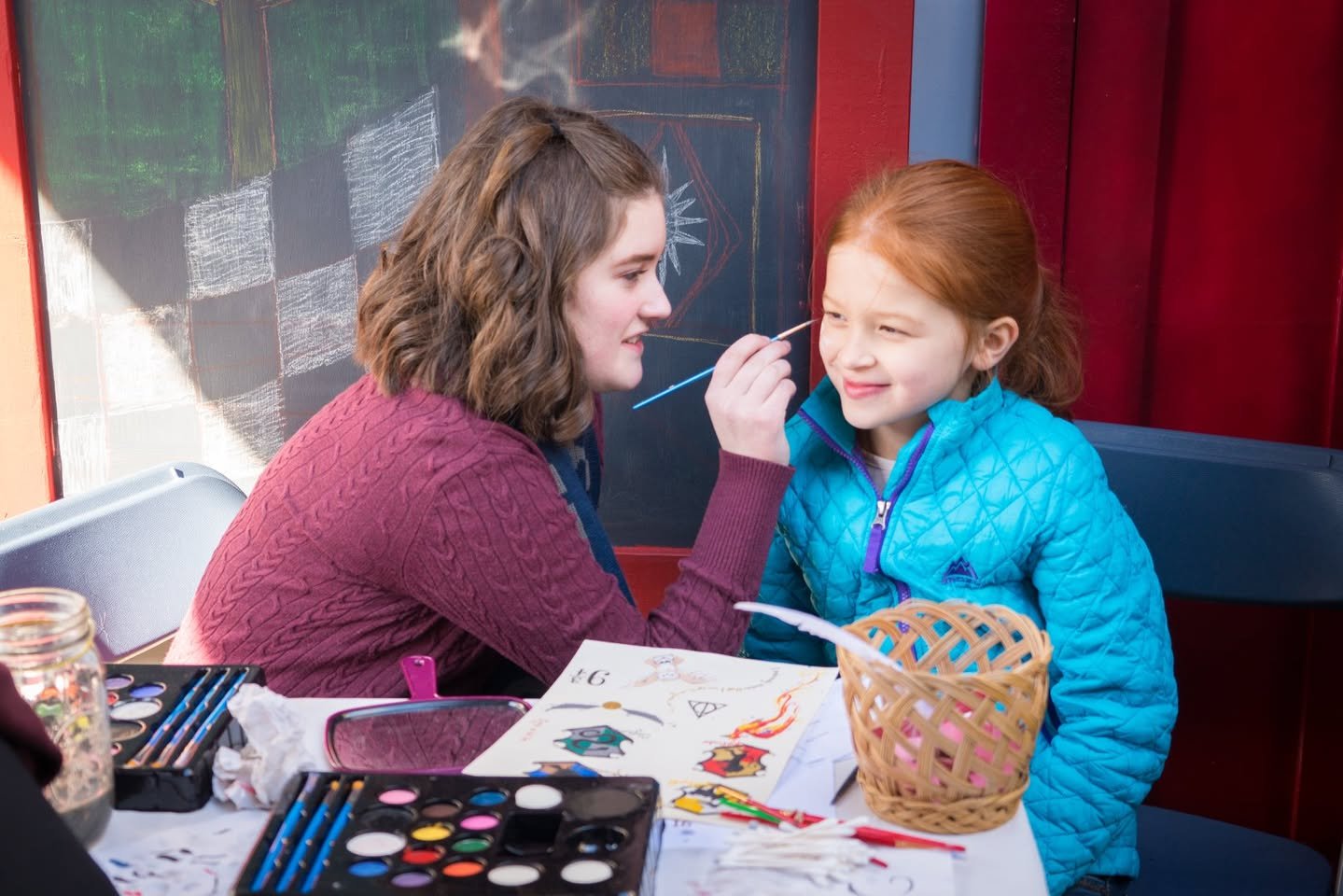 Diagon Alley A scene of face painting at an indoor craft activity. An individual is applying paint to another's face in a colorful setting.