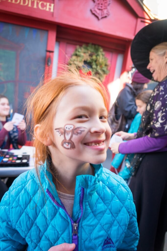 Close-up of a smiling child with Halloween face paint wearing a blue jacket at a local festival or event.
