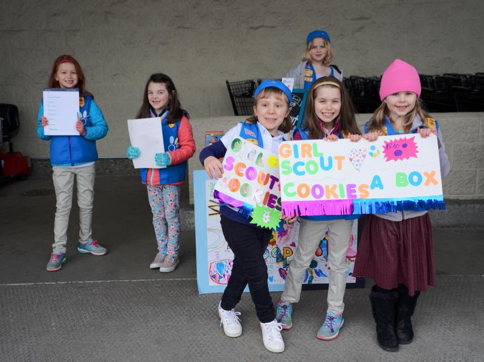 A group of Girl Scouts proudly displaying a colorful 'Girl Scout Cookies A Box' sign and certificates at an outdoor event.