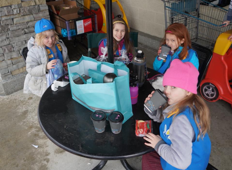 Children gather around a table outdoors, sharing a blue bag and drinks.