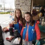 Two girls in colorful jackets sit at an outdoor dining table with drinks and snacks, surrounded by a stone pillar and shopping carts. The setting looks like a café or shop in the Pacific Northwest.