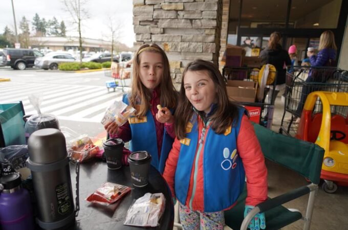 Two girls in colorful jackets sit at an outdoor dining table with drinks and snacks, surrounded by a stone pillar and shopping carts. The setting looks like a café or shop in the Pacific Northwest.