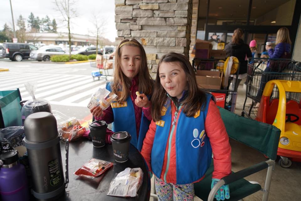 Two girls in colorful jackets sit at an outdoor dining table with drinks and snacks, surrounded by a stone pillar and shopping carts. The setting looks like a café or shop in the Pacific Northwest.