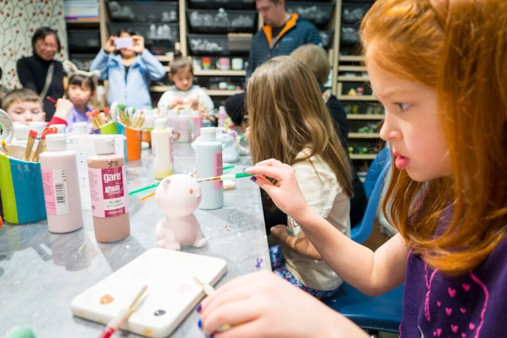 Children painting small ceramic figures at a table with watercolor supplies while parents watch.