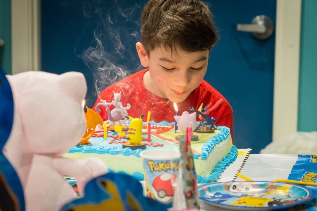A child in a red shirt exhales smoke while blowing out a birthday candle on a blue-frosted cake. The scene is filled with party-themed decorations and a cup labeled 'Poké'.