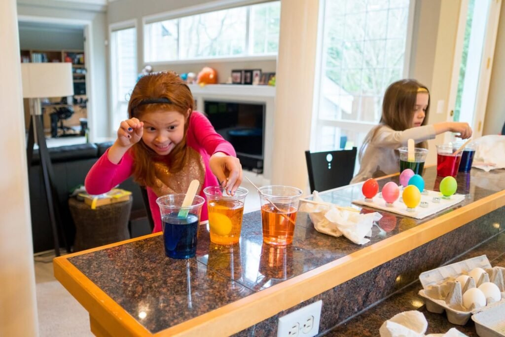 Two kids are dyeing Easter eggs with brightly colored liquids at a kitchen island.