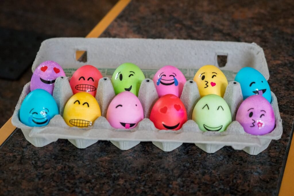 A collection of colorful plastic eggs painted with various emoji faces arranged in a grey cardboard egg carton.