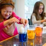 Two children performing a floating egg experiment with colorful water at a kitchen counter. One child looks delighted while observing the results.