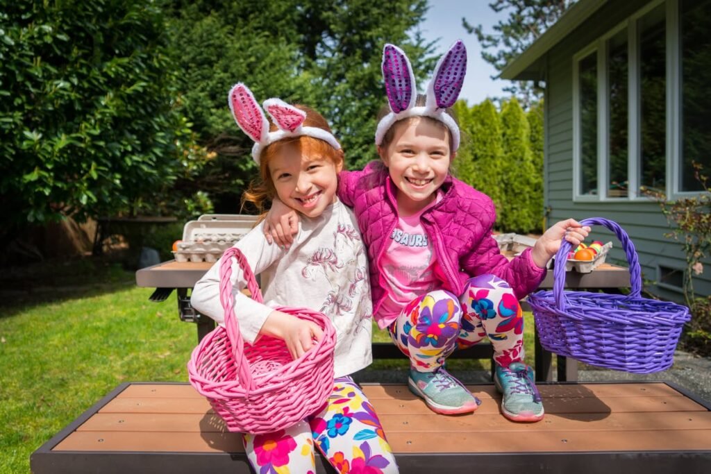 Two children wearing colorful bunny ears and costumes, sitting on a wooden bench in a sunny backyard.