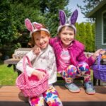 Two children wearing colorful bunny ears and costumes, sitting on a wooden bench in a sunny backyard.