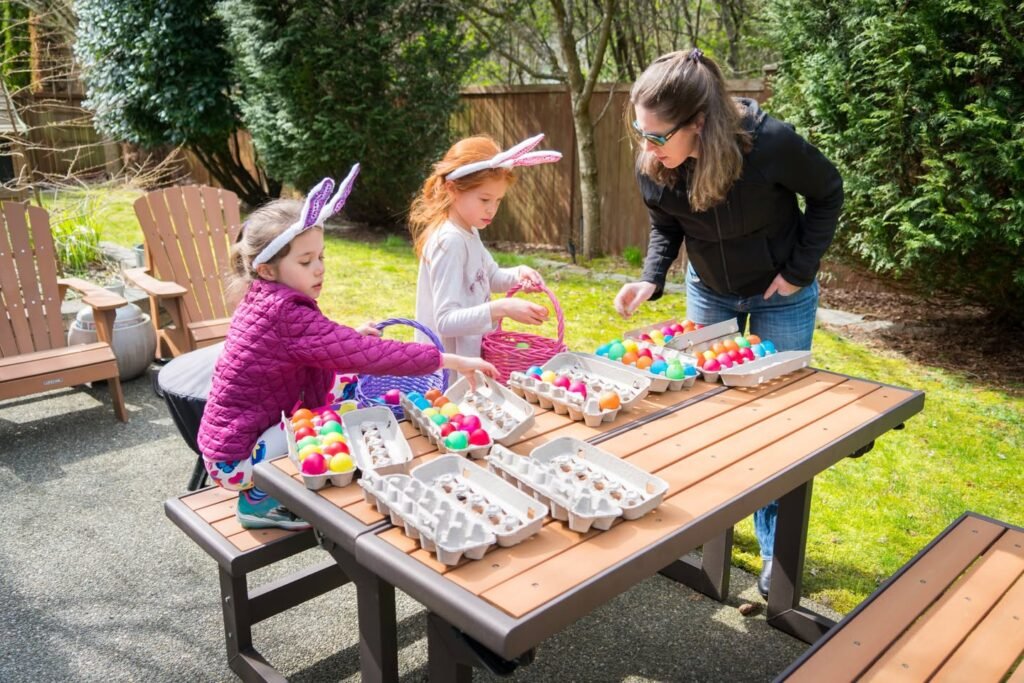 A group of people enjoys Easter egg coloring outdoors in a lush garden. The scene includes colorful eggs in cartons on a wooden table.