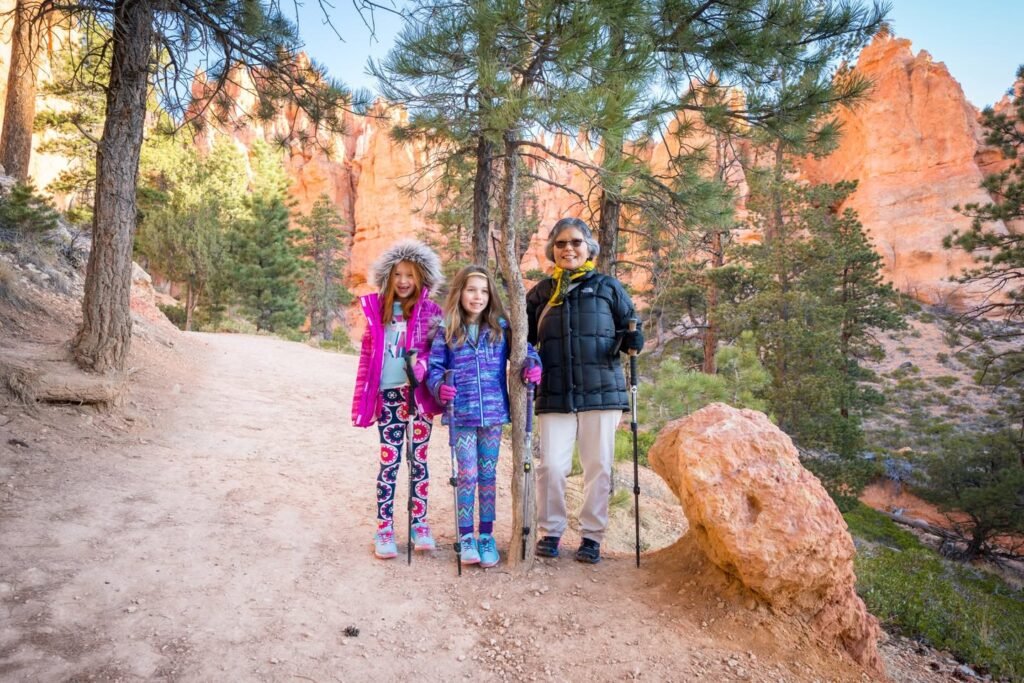 Three people equipped with hiking poles pose on a scenic trail in a pine forest with towering red rock formations in the background.