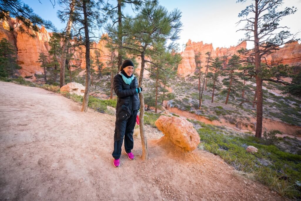 A hiker stands on a gravel trail surrounded by tall pine trees and reddish rocky cliffs in a natural landscape.