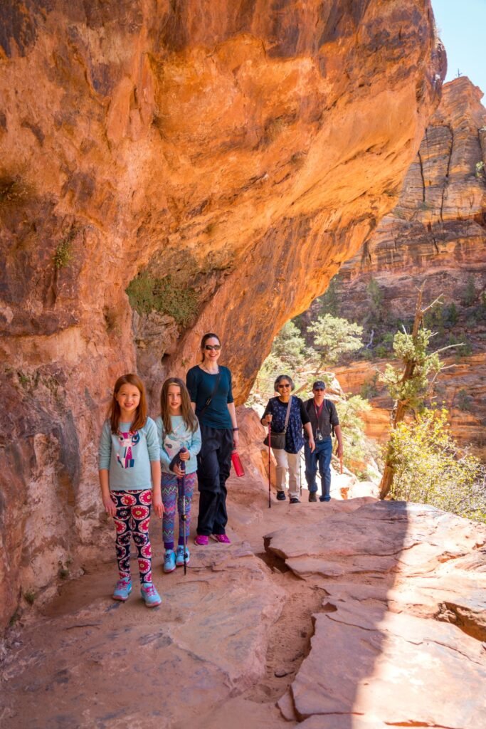 A family hiking under spectacular red rock arches in the desert, standing on a natural sandstone path with dramatic geological formations visible in the background.
