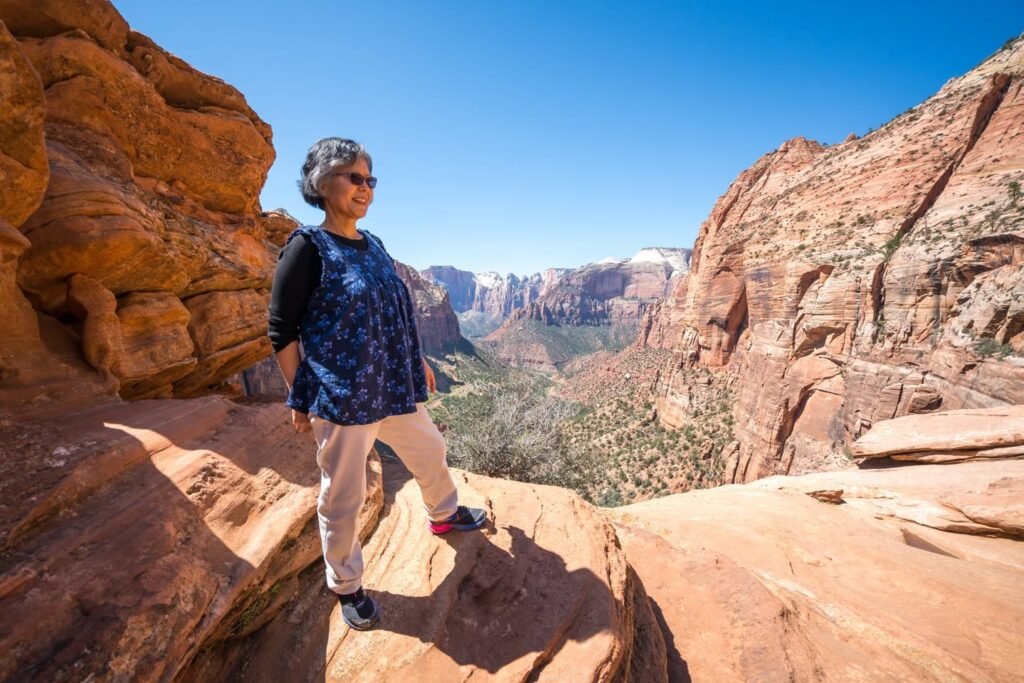 A hiker stands on a sunlit red rock ledge overlooking a vast canyon under a clear blue sky.