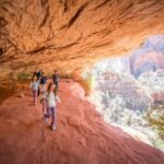 Hikers walking along a dirt path under a large red rock overhang with a sweeping view of layered canyons.