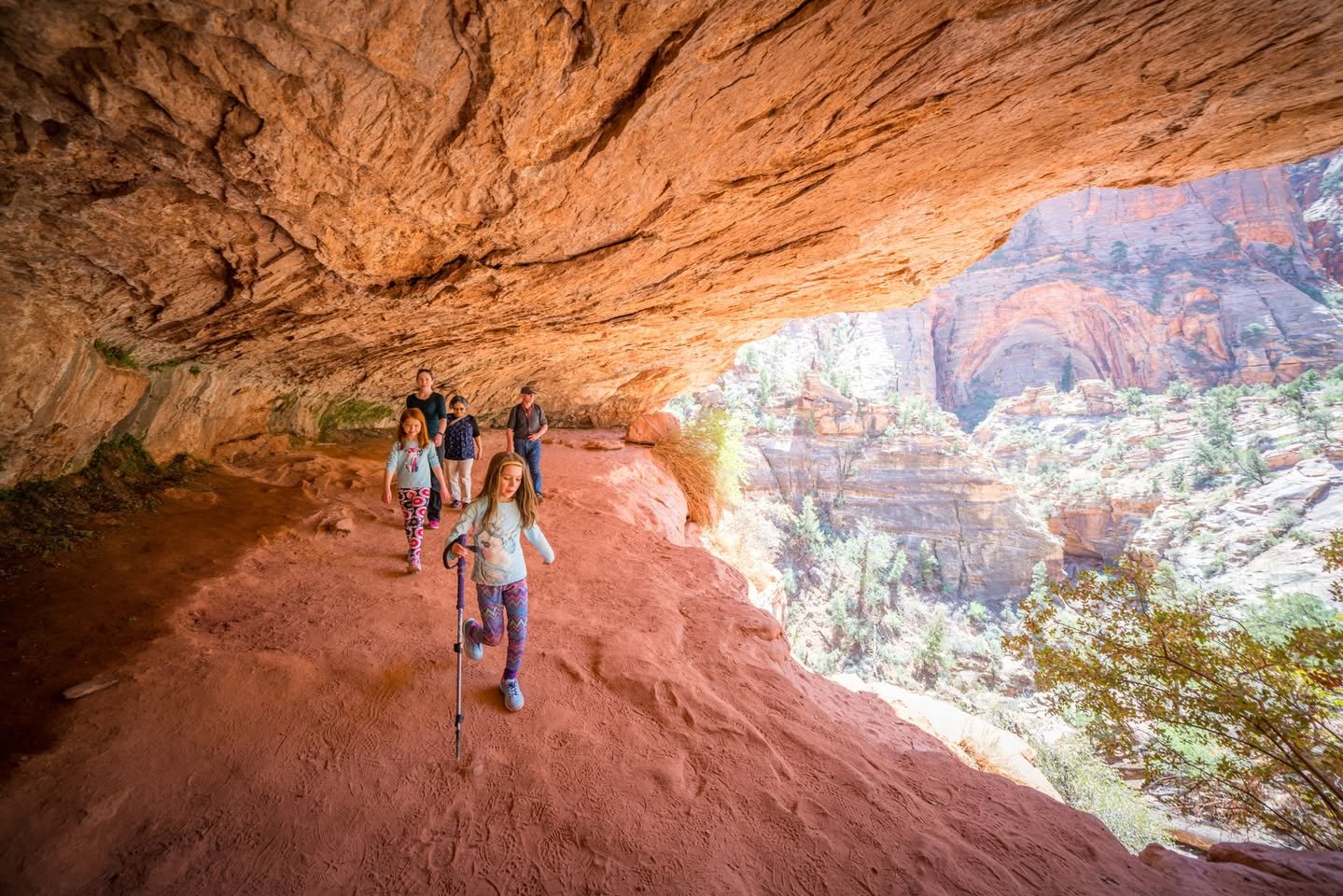 Canyon Overlook Trail Hikers walking along a dirt path under a large red rock overhang with a sweeping view of layered canyons.