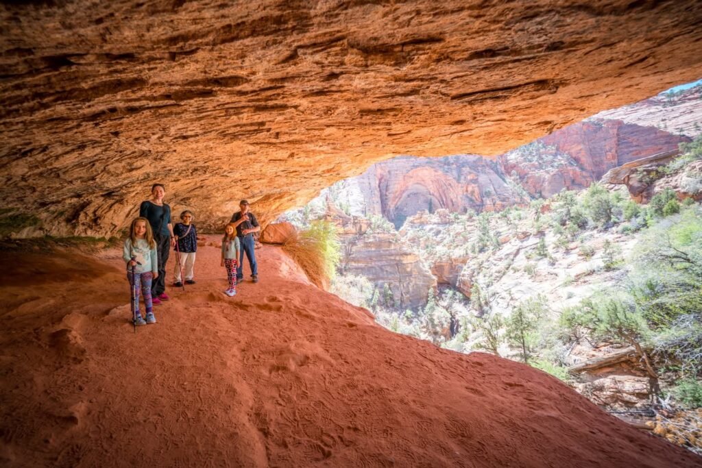 A group of hikers stands beneath a large red rock overhang in a desert canyon.