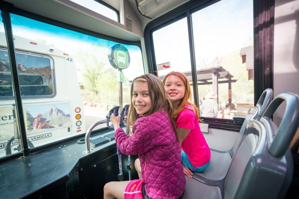 Two smiling girls sitting in the front seats of a shuttle bus, one holding the steering controls.