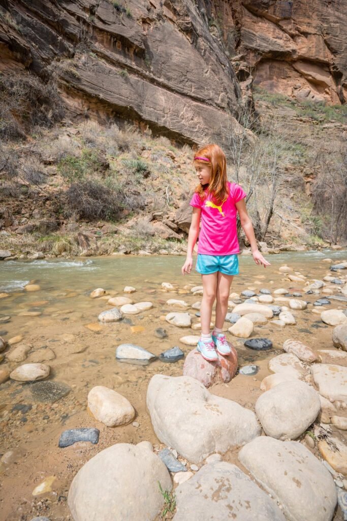 A child in bright clothing stands on large boulders in a dry riverbed beneath steep, eroded sandstone cliffs.