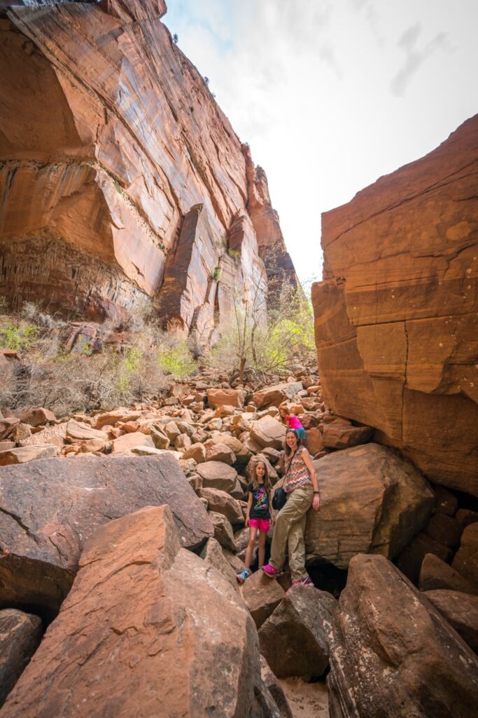 A group of hikers navigating a rocky path within towering red sandstone canyon walls under a pale sky.